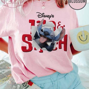 Op de afbeelding: Lichtroze T-shirt met een afbeelding van Disney's Stitch-personage dat door een papier-effect breekt. De tekst "Disney's Lilo & Stitch" staat in zwart en rood boven de afbeelding. Een witte mok met een gele smiley wordt vastgehouden.