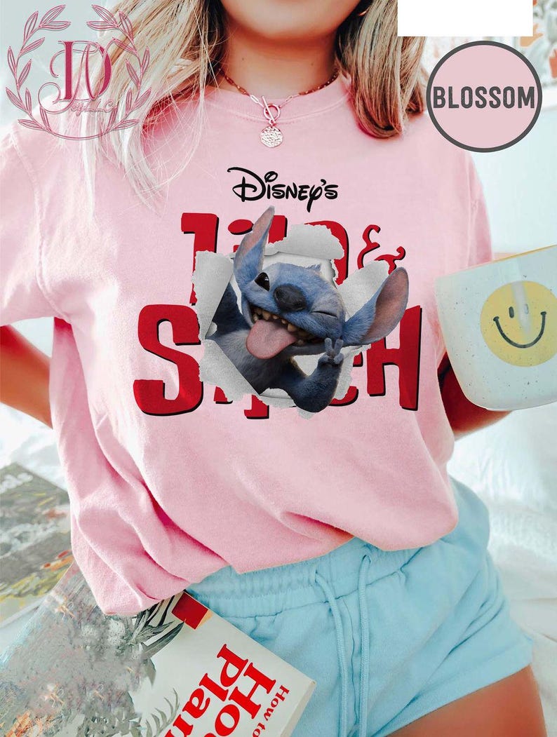 Op de afbeelding: Lichtroze T-shirt met een afbeelding van Disney's Stitch-personage dat door een papier-effect breekt. De tekst "Disney's Lilo & Stitch" staat in zwart en rood boven de afbeelding. Een witte mok met een gele smiley wordt vastgehouden.