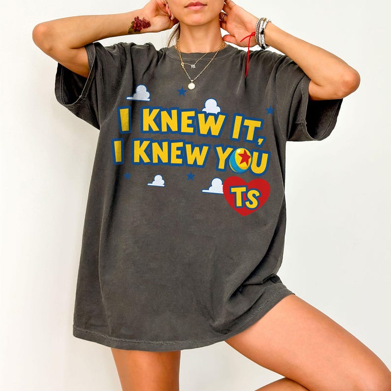 May include: A charcoal grey short-sleeve t-shirt featuring the yellow text I KNEW IT, I KNEW YOU, a colourful ball graphic, and a red heart with the letters TS. The design includes small white cloud shapes and blue stars scattered across the front.