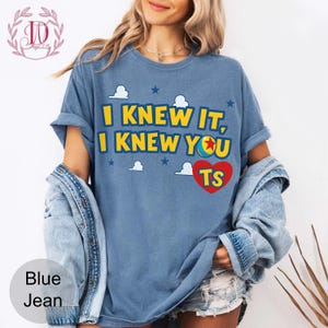 May include: A blue short-sleeve t-shirt featuring the yellow text I KNEW IT, I KNEW YOU with a colourful ball graphic replacing the letter O, a red heart with the letters TS, and white cloud and blue star graphics printed on the front.