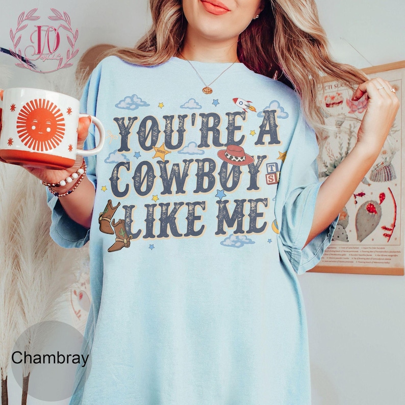 May include: A light blue t-shirt featuring the text "YOU'RE A COWBOY LIKE ME" in dark blue letters. The design includes illustrations of a brown cowboy hat, brown cowboy boots, a rocket, clouds, stars, and alphabet blocks. A white mug with a red sun graphic is held.