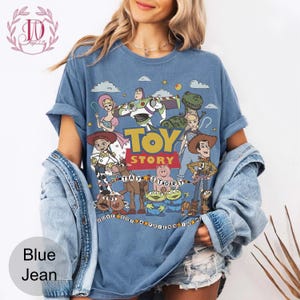 May include: A blue short-sleeve t-shirt featuring a graphic print of Toy Story characters including Woody, Buzz Lightyear, Jessie, and Rex. The design includes the text "TOY STORY" in yellow and red, along with the phrase "YOU'VE GOT A FRIEND IN ME".