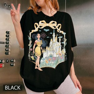 May include: Black t-shirt featuring a colorful illustration of a Native American woman in a yellow dress standing in front of a castle. The design includes flowers, fireworks, and a large yellow bow. The word "BLACK" is printed at the bottom.