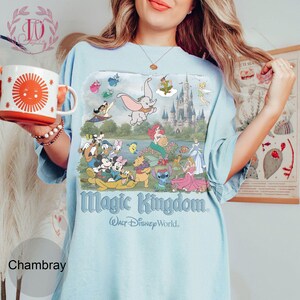 May include: A light blue t-shirt featuring a graphic print of various Disney characters including Dumbo, Mickey Mouse, and Winnie the Pooh in front of a castle. The text reads Magic Kingdom and Walt Disney World in a stylized blue font.