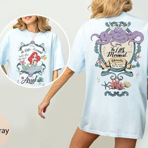 May include: Light blue t-shirt featuring a graphic from Disney's The Little Mermaid. The front design includes Ariel, while the back showcases Ursula. The shirt has short sleeves and a relaxed fit, perfect for casual wear.