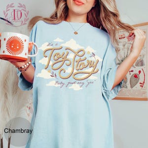 May include: A light blue t-shirt featuring a graphic print of the text 'it's a Toy Story' written in a rope-like font, surrounded by white clouds and small floral accents. The text 'Baby, just say "yes"' is printed below in a cursive script.