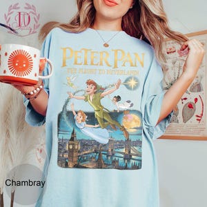 May include: Pale blue t-shirt with a Peter Pan graphic showing characters flying over London. The text "PETER PAN THE FLIGHT TO NEVERLAND!" is printed in gold. The shirt is a chambray colour.