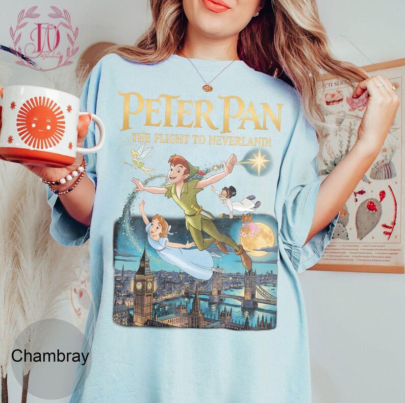 May include: Pale blue t-shirt with a Peter Pan graphic showing characters flying over London. The text "PETER PAN THE FLIGHT TO NEVERLAND!" is printed in gold. The shirt is a chambray colour.
