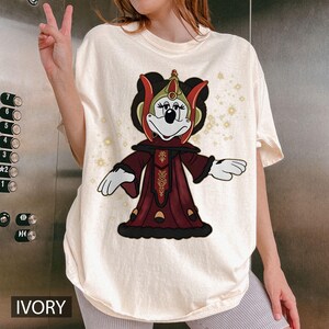 Puede incluir: Camiseta de color marfil con un personaje de dibujos animados con un disfraz rojo y dorado. El personaje tiene un tocado negro y est&aacute; sobre un fondo de peque&ntilde;as estrellas doradas brillantes. La palabra "IVORY" est&aacute; impresa en la parte inferior.