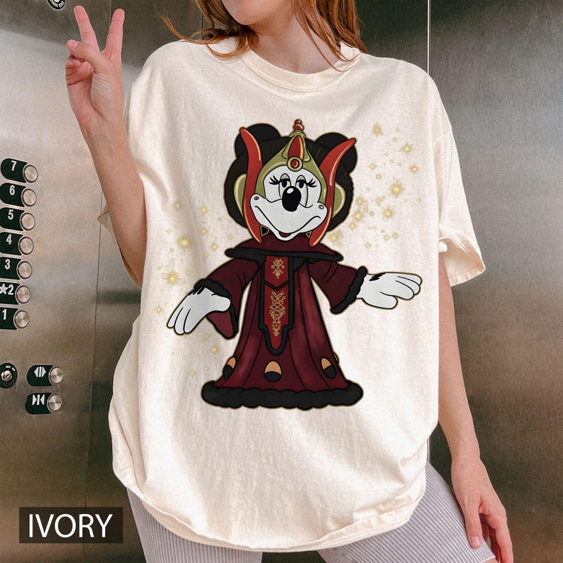 Puede incluir: Camiseta de color marfil con un personaje de dibujos animados con un disfraz rojo y dorado. El personaje tiene un tocado negro y est&aacute; sobre un fondo de peque&ntilde;as estrellas doradas brillantes. La palabra "IVORY" est&aacute; impresa en la parte inferior.