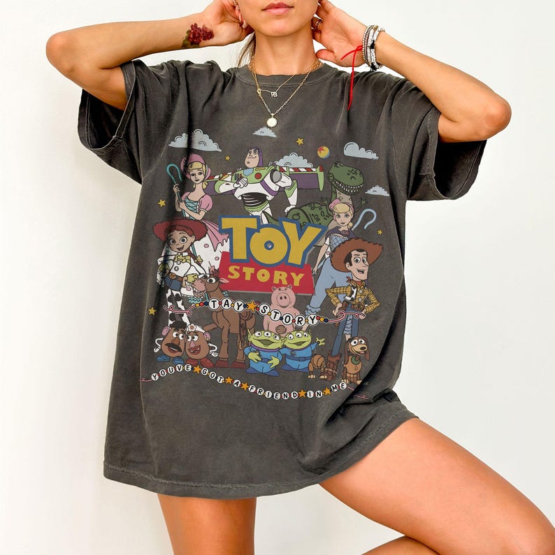 May include: A charcoal gray short-sleeve t-shirt featuring a large graphic print of Toy Story characters including Woody, Buzz Lightyear, Jessie, and others. The text Toy Story is printed in large yellow and blue letters with the phrase You've Got A Friend In Me.