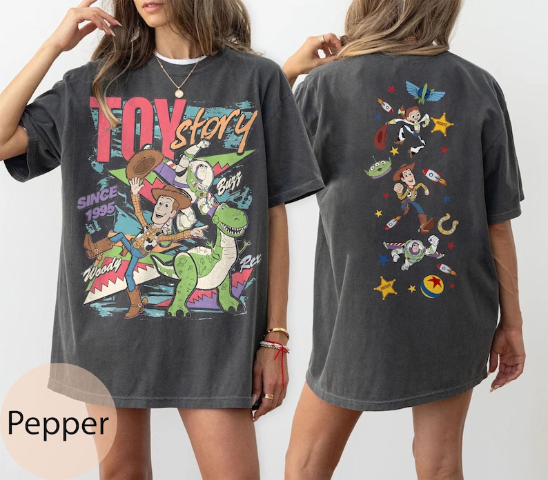 May include: Dark grey t-shirt featuring the Toy Story logo and characters Woody, Buzz Lightyear, and Rex on the front. The back of the shirt displays additional Toy Story characters. The shirt has a vintage, distressed look.