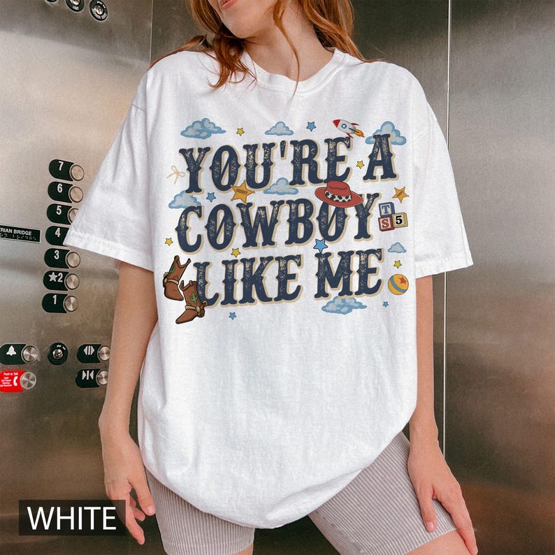 May include: A white short-sleeve t-shirt featuring the text "YOU'RE A COWBOY LIKE ME" in dark blue letters. The design includes illustrations of brown cowboy boots, a red cowboy hat, a rocket, a colorful ball, and small blue clouds and stars.