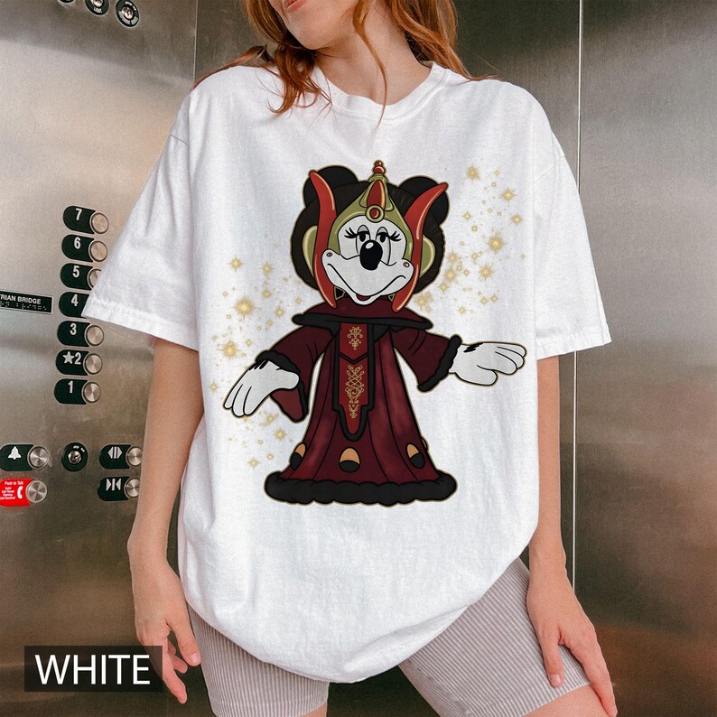 Puede incluir: Camiseta blanca con un personaje de dibujos animados con un atuendo burdeos y dorado. El personaje tiene un tocado negro y est&aacute; sobre un fondo de destellos dorados. La palabra "WHITE" est&aacute; impresa en la parte inferior.