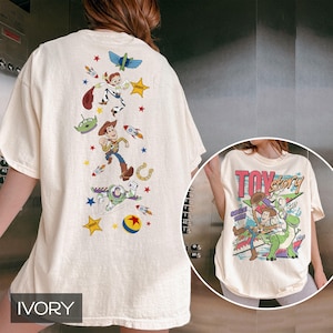 May include: Ivory t-shirt with a colourful cartoon design and the text "TOY STORY" with "SINCE 1995". The back of the shirt features Woody, Buzz Lightyear, Jessie, and other characters from the film.