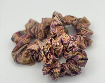 May include: Several patterned scrunchies in tan, purple, and yellow. The fabric has a geometric design with triangles and lines. These hair accessories are a stylish addition to any hairstyle.