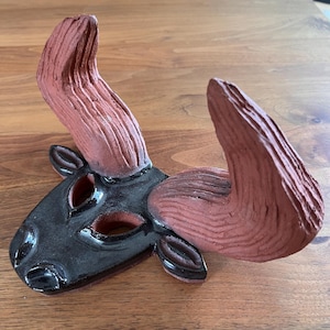 May include: A ceramic wall mask shaped like a bull head with large, textured, reddish-brown horns and a smooth, glossy black face. The mask features cut-out eye sockets and nostrils, with a distinct contrast between the matte clay horns and the glazed black finish of the face.