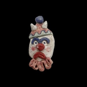 May include: A handmade ceramic clown face with a white base, blue eyes, and a red nose and mouth. The clown wears a pink and blue hat and a pink ruffled collar. Black speckles add detail to the face.