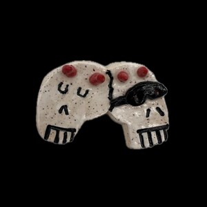 May include: A ceramic pin featuring two conjoined white skulls with black speckled details. The left skull has red accents and black facial features, while the right skull wears a black eye patch and has red accents, all set against a black background.