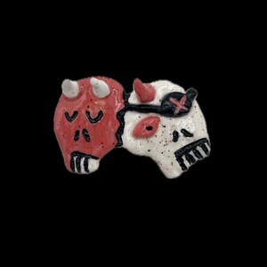 May include: A ceramic sculpture featuring two conjoined halves: a red devil face and a white skull. The devil half has horns and closed eyes, while the skull has an eye patch and a stitched mouth. Black details and speckles.