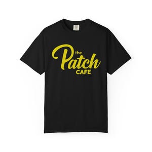 May include: Black t-shirt featuring the yellow text "the Patch CAFE" in a cursive font. The shirt has a crew neck design. The text is centered on the front of the shirt.