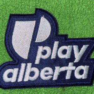 Könnte beinhalten: Ein dunkelblauer gestickter Aufnäher mit einem weißen stilisierten Schildsymbol und dem Text play alberta in weißen Kleinbuchstaben. Der Aufnäher hat eine markante geometrische Form mit einem dicken dunkelblauen Rand und einer strukturierten Stoffoberfläche.