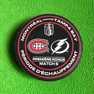 May include: A black hockey puck featuring the text MONTRÉAL CONTRE TAMPA BAY, PREMIÈRE RONDE, and MATCH 6. The center displays the Montreal Canadiens logo, a Tampa Bay Lightning logo, and a small Stanley Cup graphic with the years 2026.