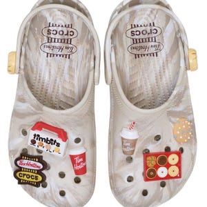 May include: A pair of beige marbled clogs featuring a swivel heel strap and various Tim Hortons themed charms. The footbed displays the Crocs and Tim Hortons logos. The charms include a Timbits box, coffee cups, a donut, and a branded logo patch.