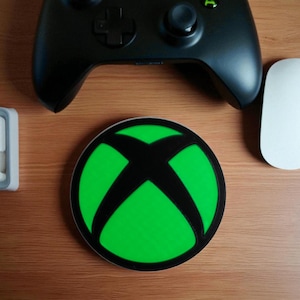 May include: A round, green and black Xbox logo coaster sits on a wooden surface, with a black Xbox controller and a white mouse nearby. The coaster features the iconic Xbox logo in black over a bright green background.
