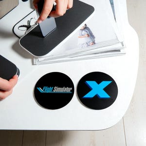 May include: A white table with two black coasters and a stack of photos. One coaster has the text "Flight Simulator" and the other has a blue "X". A person is holding a black object.