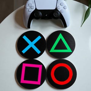 May include: A white game controller rests above four black circular coasters. Each coaster features a different colored PlayStation symbol: blue X, green triangle, pink square, and red circle. The coasters are designed for gaming enthusiasts.