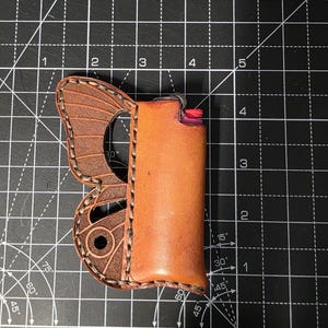 Puede incluir: Un estuche de cuero marr&oacute;n para encendedor con dise&ntilde;o de ala de mariposa. El estuche contiene un encendedor. El ala de mariposa est&aacute; en el lado izquierdo del estuche. El estuche es de cuero marr&oacute;n.