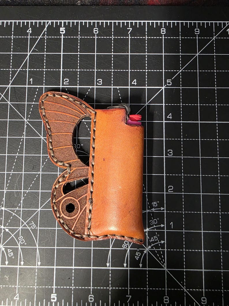 Puede incluir: Un estuche de cuero marr&oacute;n para encendedor con dise&ntilde;o de ala de mariposa. El estuche contiene un encendedor. El ala de mariposa est&aacute; en el lado izquierdo del estuche. El estuche es de cuero marr&oacute;n.