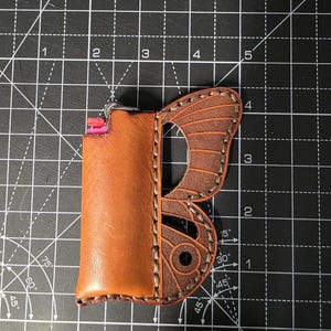 Puede incluir: Una funda de cuero marr&oacute;n para encendedor con dise&ntilde;o de ala de mariposa. La funda est&aacute; cosida con hilo en contraste y tiene un llavero de metal. El encendedor es visible dentro de la funda. El ala de mariposa es de color marr&oacute;n oscuro.