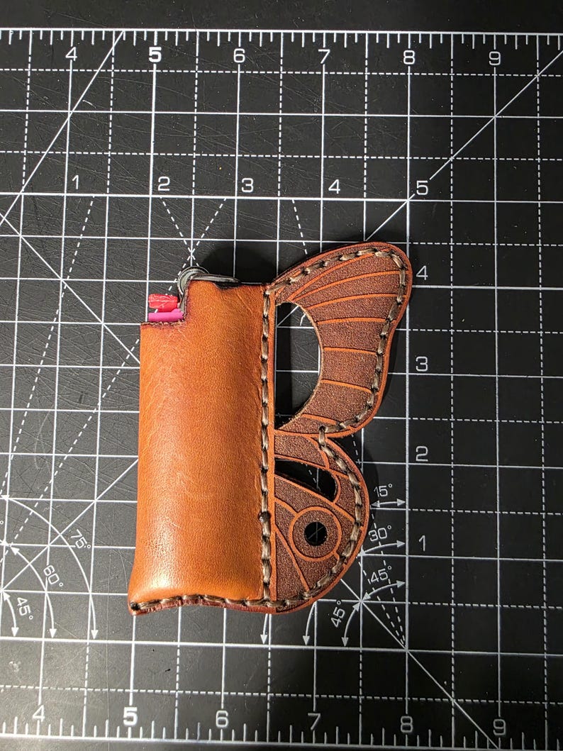 Puede incluir: Una funda de cuero marr&oacute;n para encendedor con dise&ntilde;o de ala de mariposa. La funda est&aacute; cosida con hilo en contraste y tiene un llavero de metal. El encendedor es visible dentro de la funda. El ala de mariposa es de color marr&oacute;n oscuro.