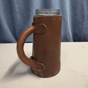 May include: A clear glass jar with a brown leather sleeve and handle. The leather sleeve is stitched and has decorative metal accents at the base. The handle is curved and attached to the sleeve with leather straps.