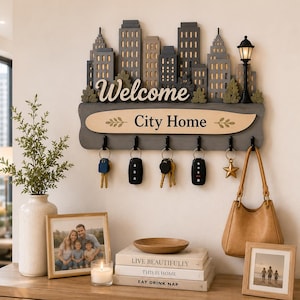 May include: A wall-mounted wooden key holder featuring a layered city skyline silhouette, a street lamp, and the text "Welcome City Home". It includes six black metal hooks holding various keys and a star charm, with a tan wooden accent panel.