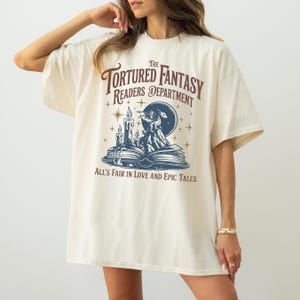 以下が含まれることがあります： 「The Tortured Fantasy Readers Department」と書かれたクリーム色のTシャツ。デザインは開いた本、ろうそく、ドラゴンに似た生き物を特徴としています。「All's Fair in Love and Epic Tales」というテキストも含まれています。