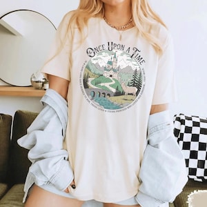 Puede incluir: Camiseta color crema con un gr&aacute;fico circular. El gr&aacute;fico muestra un castillo, monta&ntilde;as, un ciervo y un r&iacute;o, con el texto "Once Upon a Time". La camiseta tiene mangas cortas y un corte relajado.