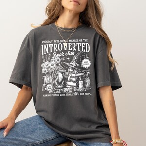 May include: Dark gray t-shirt with a graphic that reads "PROUDLY ANTI-SOCIAL MEMBER OF THE INTROVERTED Book club." The graphic features a witch hat, books, candles, a dragon, and flowers. The text "MAKING FRIENDS WITH CHARACTERS, NOT PEOPLE" is also included.
