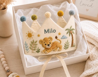 May include: A cream-colored felt crown featuring an embroidered lion, a palm tree, a sun, and the name Milo. The crown is topped with five small, multi-colored pom-poms and includes two long fabric ties at the base for securing the item.