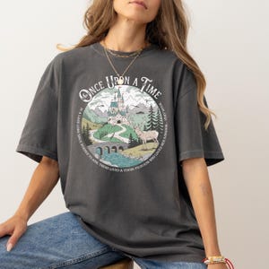 Puede incluir: Camiseta gris oscuro con un dise&ntilde;o gr&aacute;fico circular que presenta un castillo, monta&ntilde;as y un ciervo. El texto "ONCE UPON A TIME" est&aacute; en la parte superior, con texto adicional alrededor. La camiseta tiene mangas cortas y un corte holgado.
