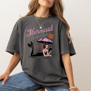 May include: A dark gray t-shirt with a graphic print of a mermaid reading a book under an umbrella. The text "The Mermaid Book Club" is printed above the graphic.