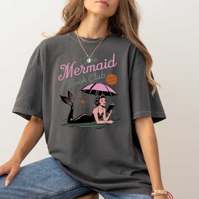 May include: A dark gray t-shirt with a graphic print of a mermaid reading a book under an umbrella. The text "The Mermaid Book Club" is printed above the graphic.