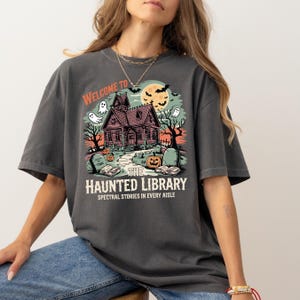 May include: Dark gray t-shirt with a Halloween-themed graphic. The design features a haunted house, ghosts, bats, and a pumpkin, with the text "WELCOME TO THE HAUNTED LIBRARY" and "SPECTRAL STORIES IN EVERY AISLE".