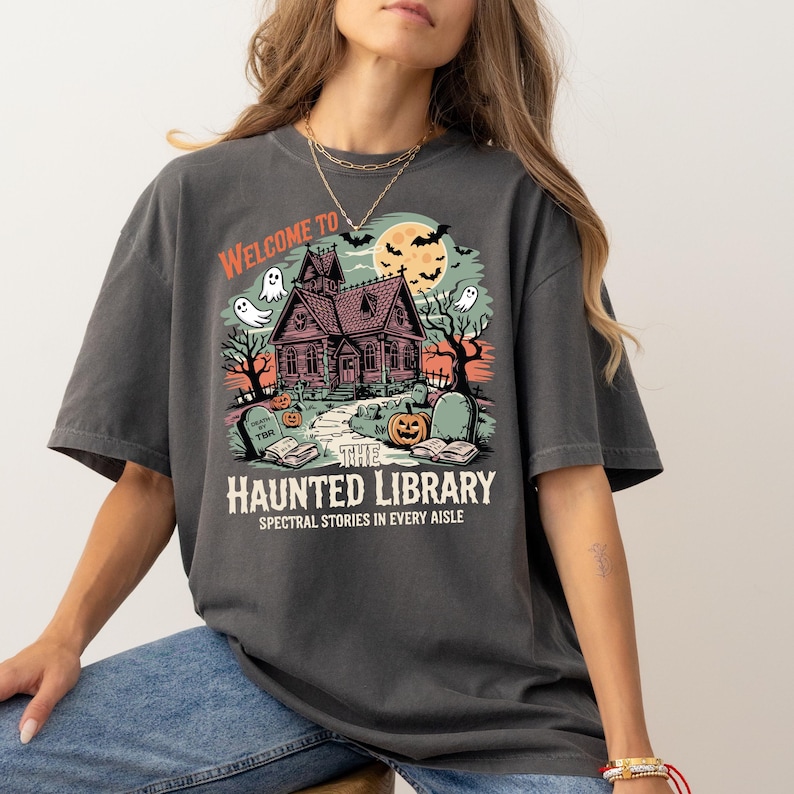 May include: Dark gray t-shirt with a Halloween-themed graphic. The design features a haunted house, ghosts, bats, and a pumpkin, with the text "WELCOME TO THE HAUNTED LIBRARY" and "SPECTRAL STORIES IN EVERY AISLE".