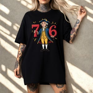 May include: Black t-shirt with a cartoon illustration of a man in colonial clothing holding the numbers "76". The man wears a blue coat, yellow vest, and white trousers. The numbers are red with yellow accents. The design celebrates a historical theme.