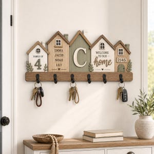 Könnte beinhalten: Ein hölzerner Wand-Schlüsselhalter mit fünf schwarzen Metallhaken und einem dekorativen Design aus fünf Häusern. Die Häuser zeigen den Text FAMILY OF 4, die Namen Emma, Jacob, Noah, Lily, den Buchstaben C, WELCOME TO OUR home und die Zahl 2145.