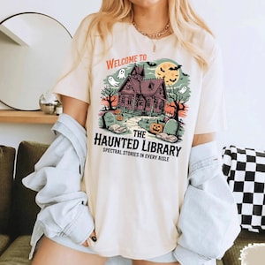 May include: Cream-colored t-shirt featuring a Halloween-themed graphic. The design includes a haunted house, ghosts, pumpkins, and the text "Welcome to The Haunted Library: Spectral Stories in Every Aisle."