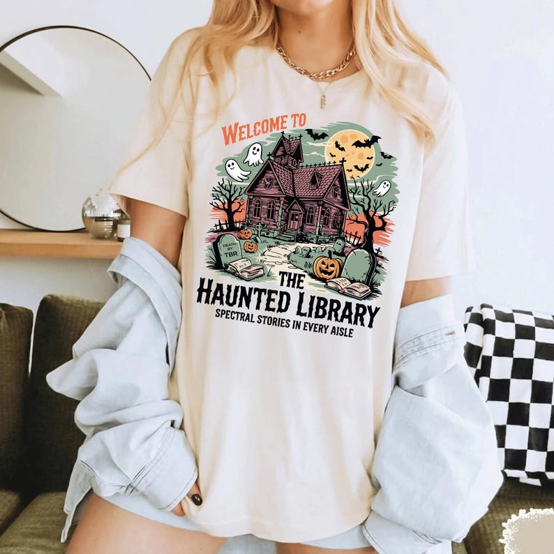 May include: Cream-colored t-shirt featuring a Halloween-themed graphic. The design includes a haunted house, ghosts, pumpkins, and the text "Welcome to The Haunted Library: Spectral Stories in Every Aisle."
